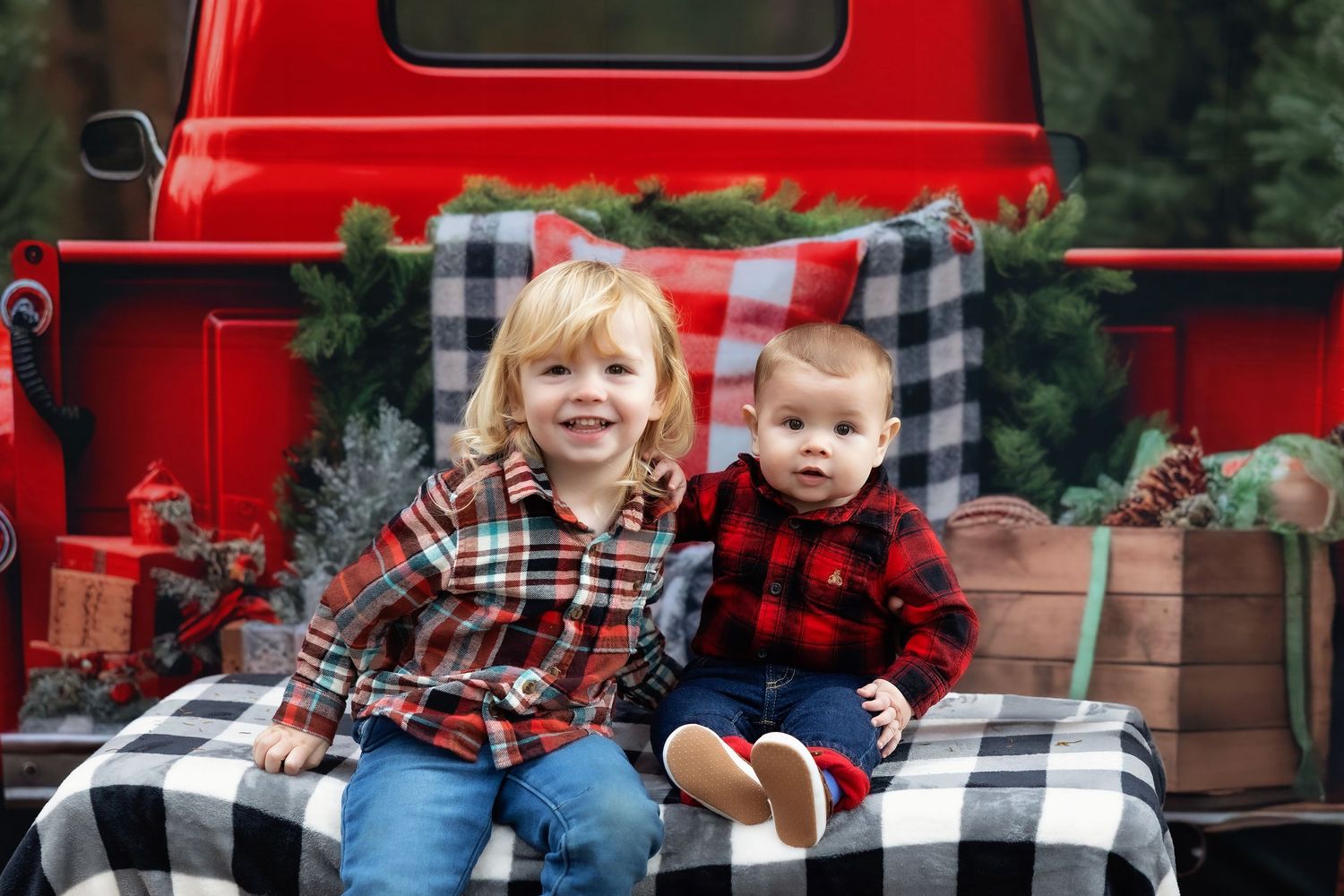 Two children in plaid shirts sit on a red vintage truck tailgate with Christmas greenery and gifts for a holiday photo.