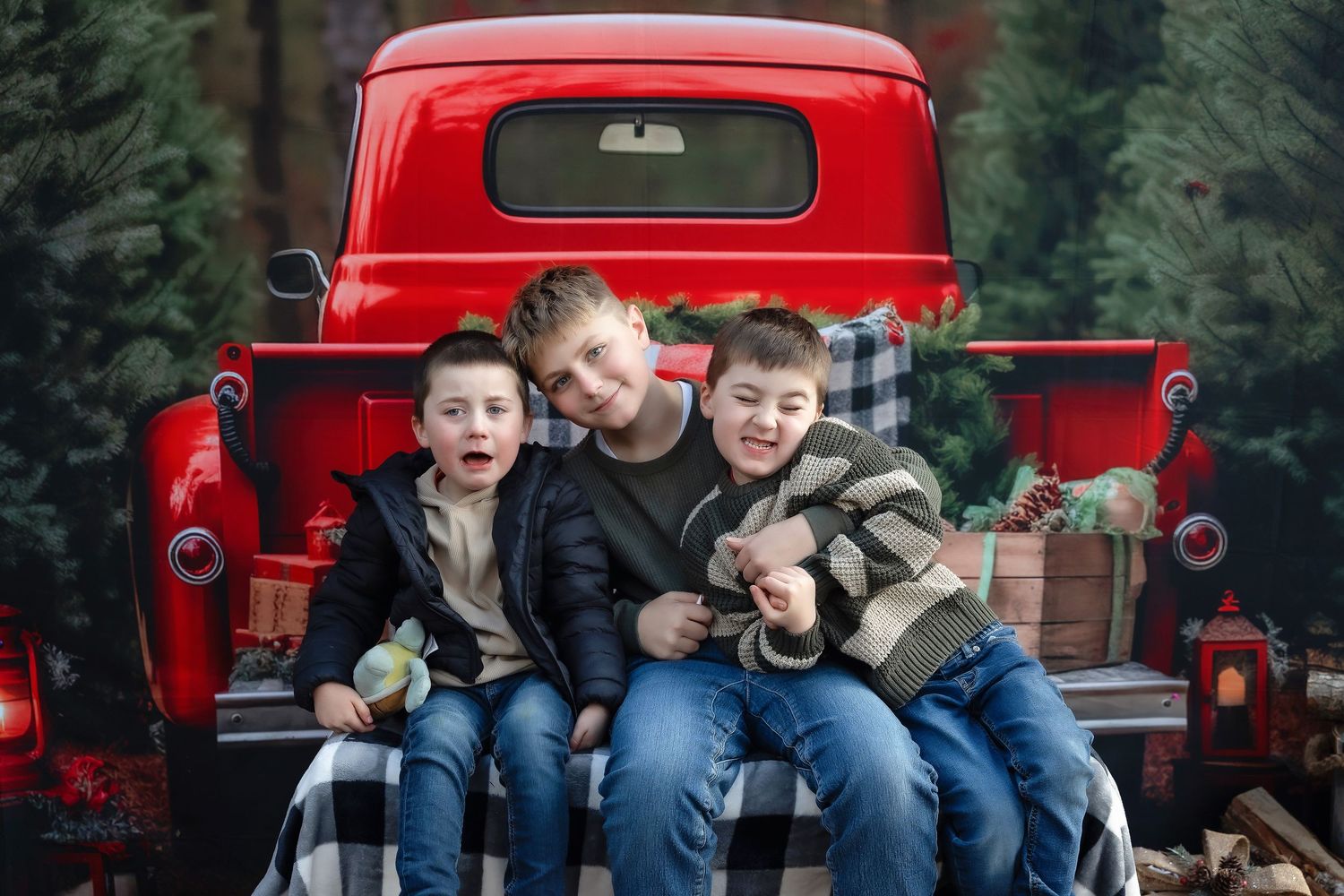 Three children sit on a red vintage truck tailgate surrounded by Christmas trees and holiday decorations.