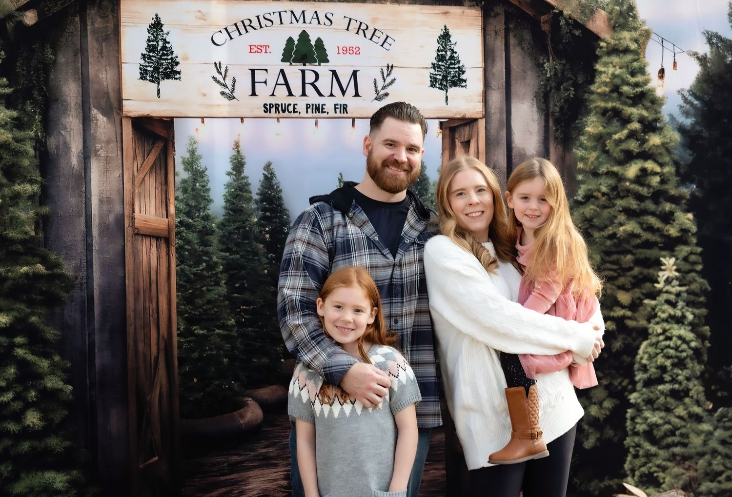 Family of four posing at a Christmas Tree Farm sign, surrounded by festive pine trees in a holiday photo.