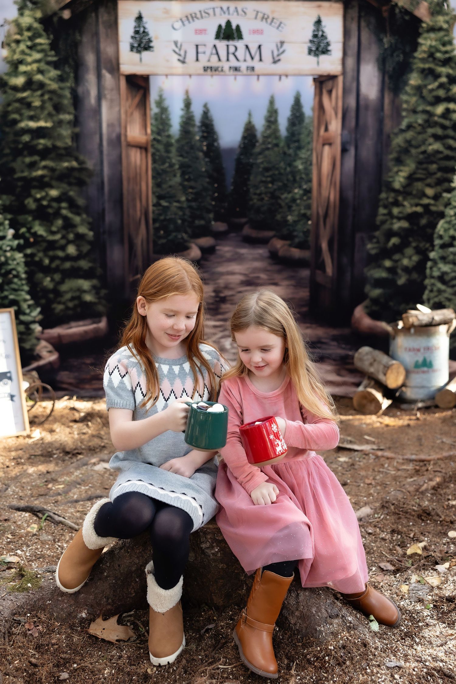 Two girls sitting on a log at a Christmas Tree Farm backdrop, holding festive mugs in holiday outfits.
