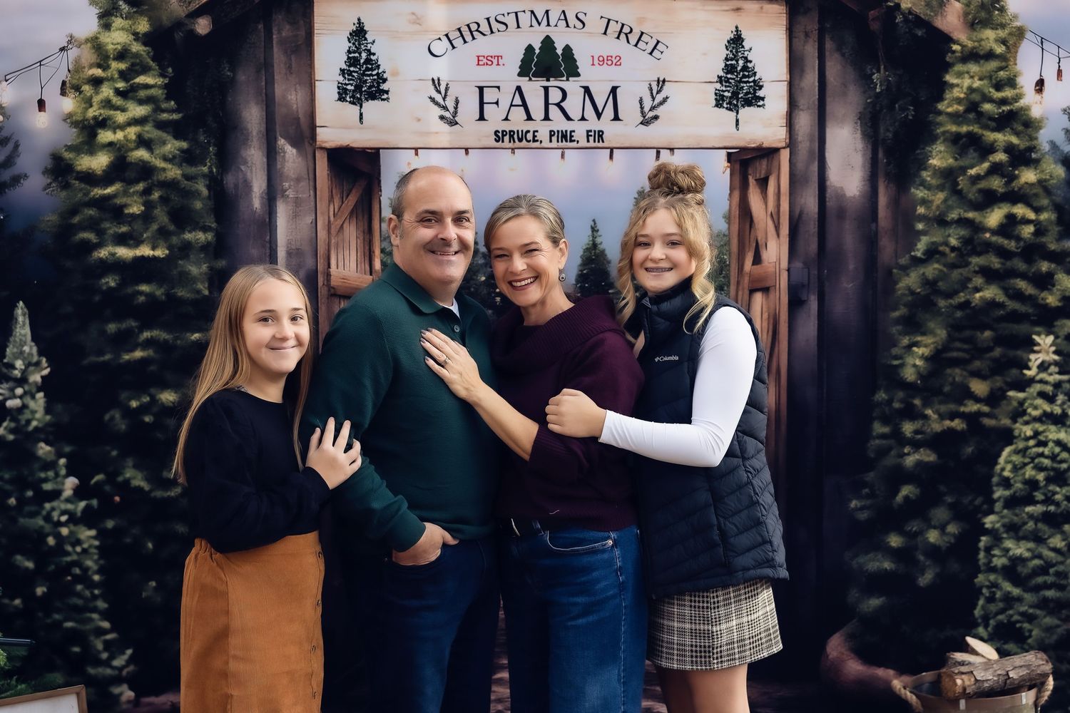 Family of four smiling together at a Christmas Tree Farm holiday photo backdrop with festive greenery.