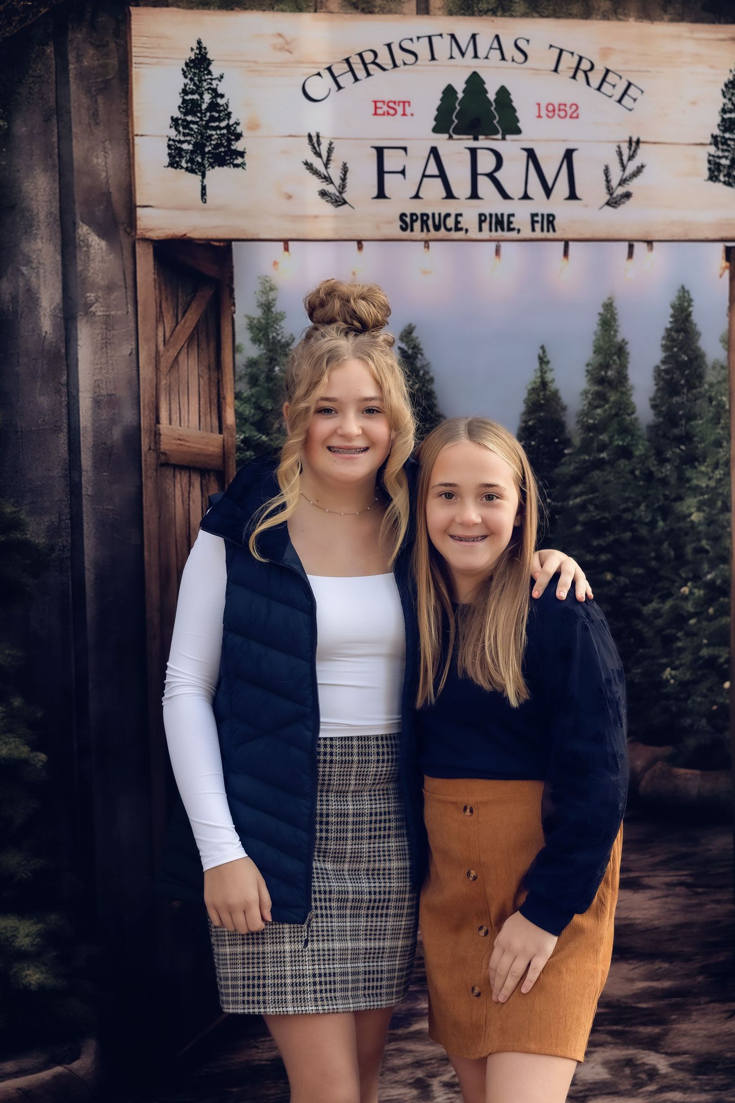Two teenage girls smiling together in front of a Christmas Tree Farm sign with pine trees in the background.