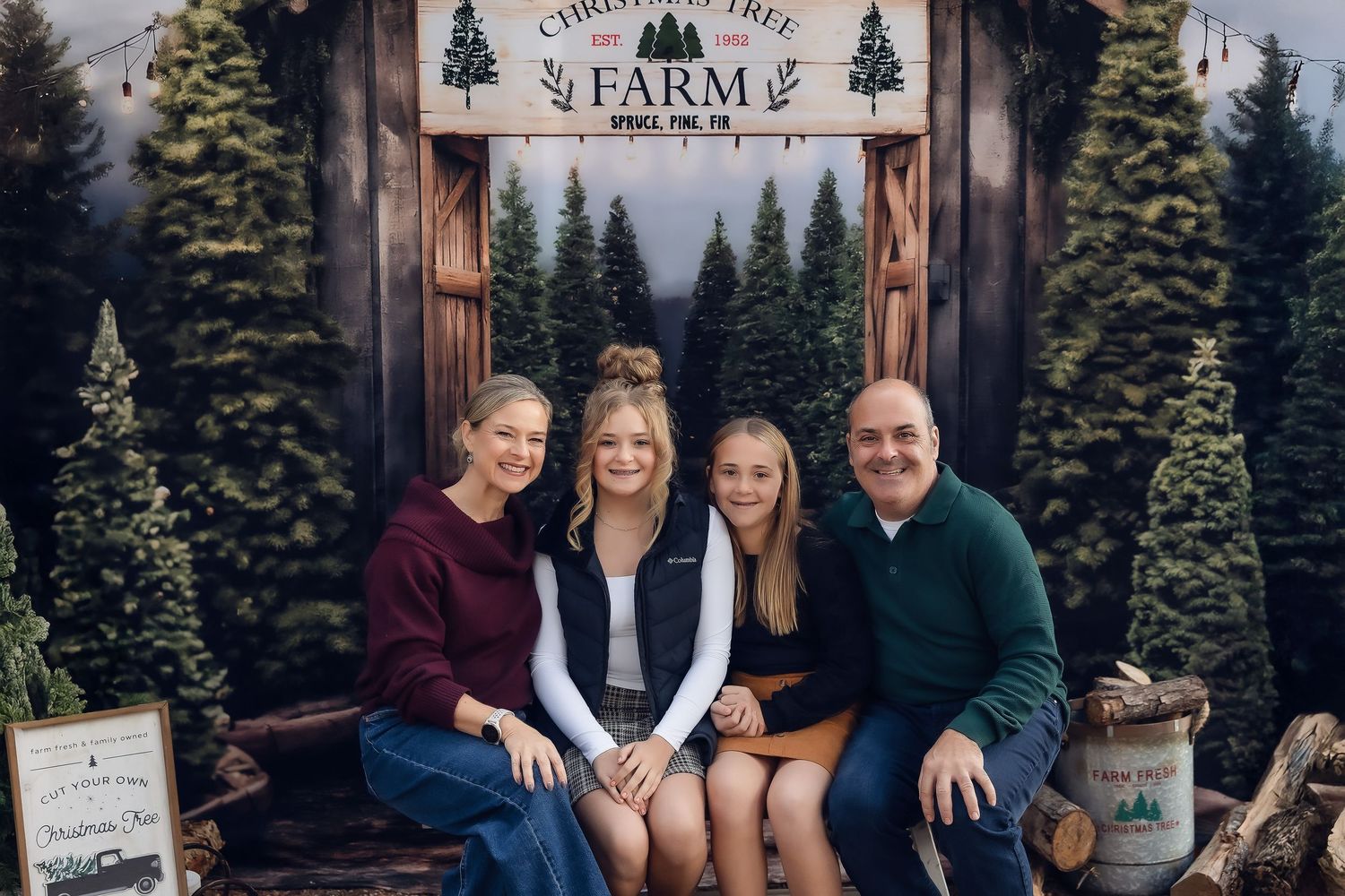 Family of four smiling together at a Christmas tree farm holiday photo session with festive backdrop.