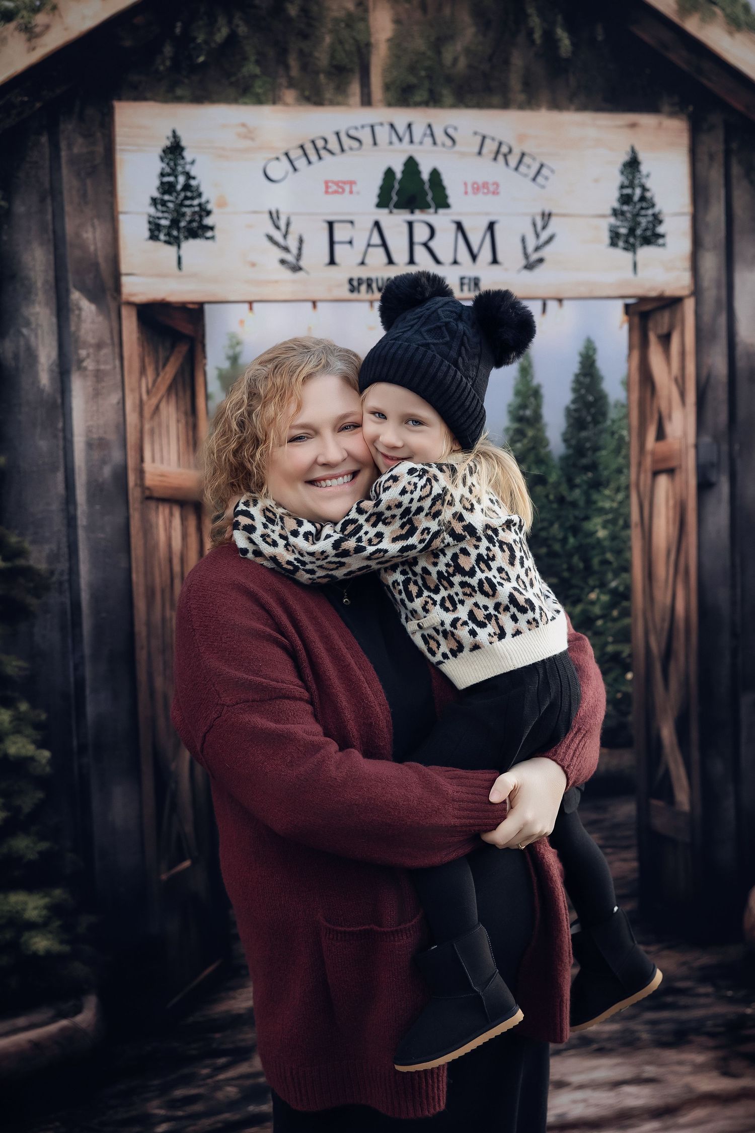 Woman holding child at Christmas Tree Farm sign, both wearing festive winter clothes, smiling warmly outdoors.
