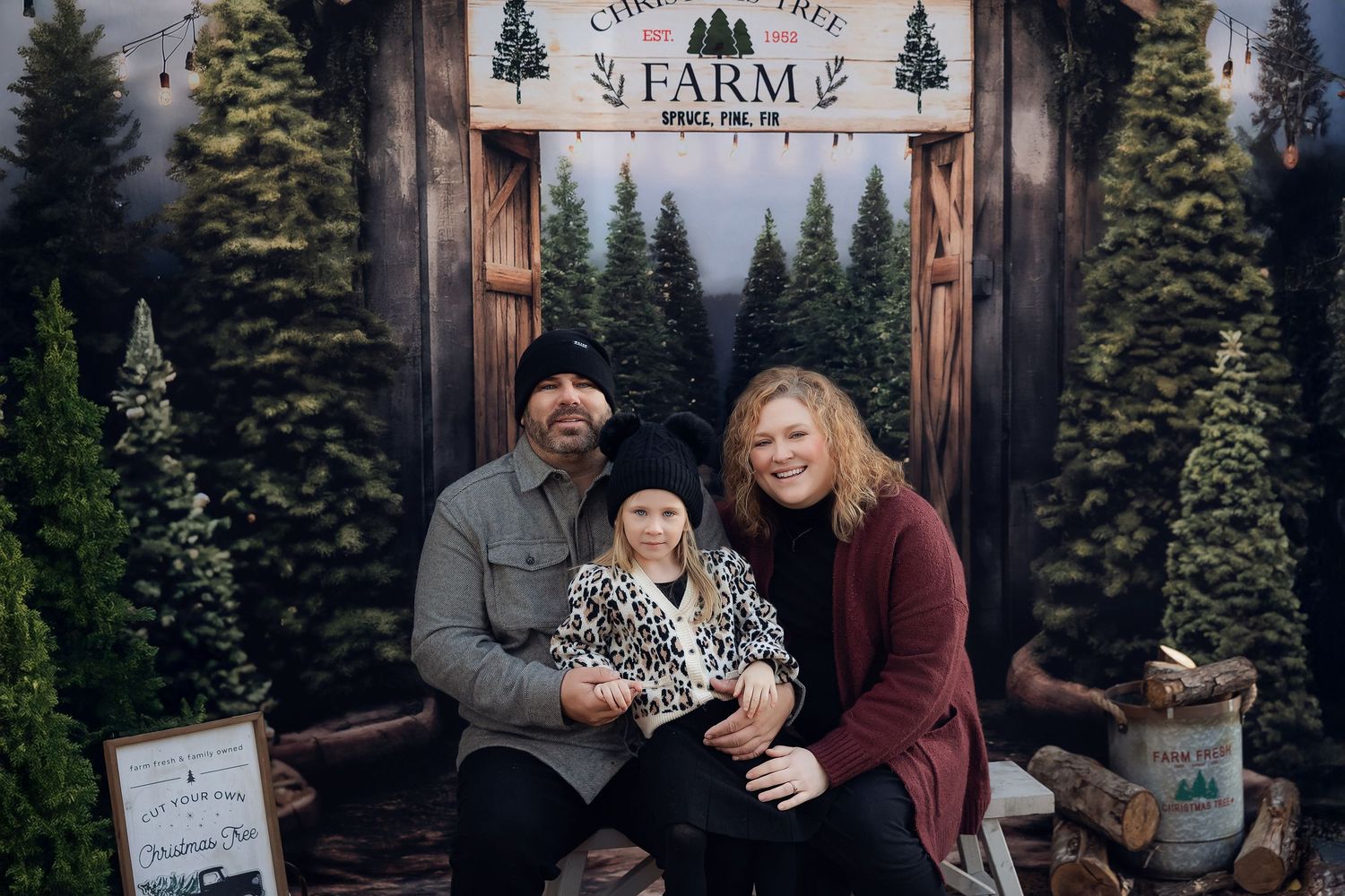 Family of three posing at a Christmas tree farm backdrop with evergreen trees and rustic wooden sign.