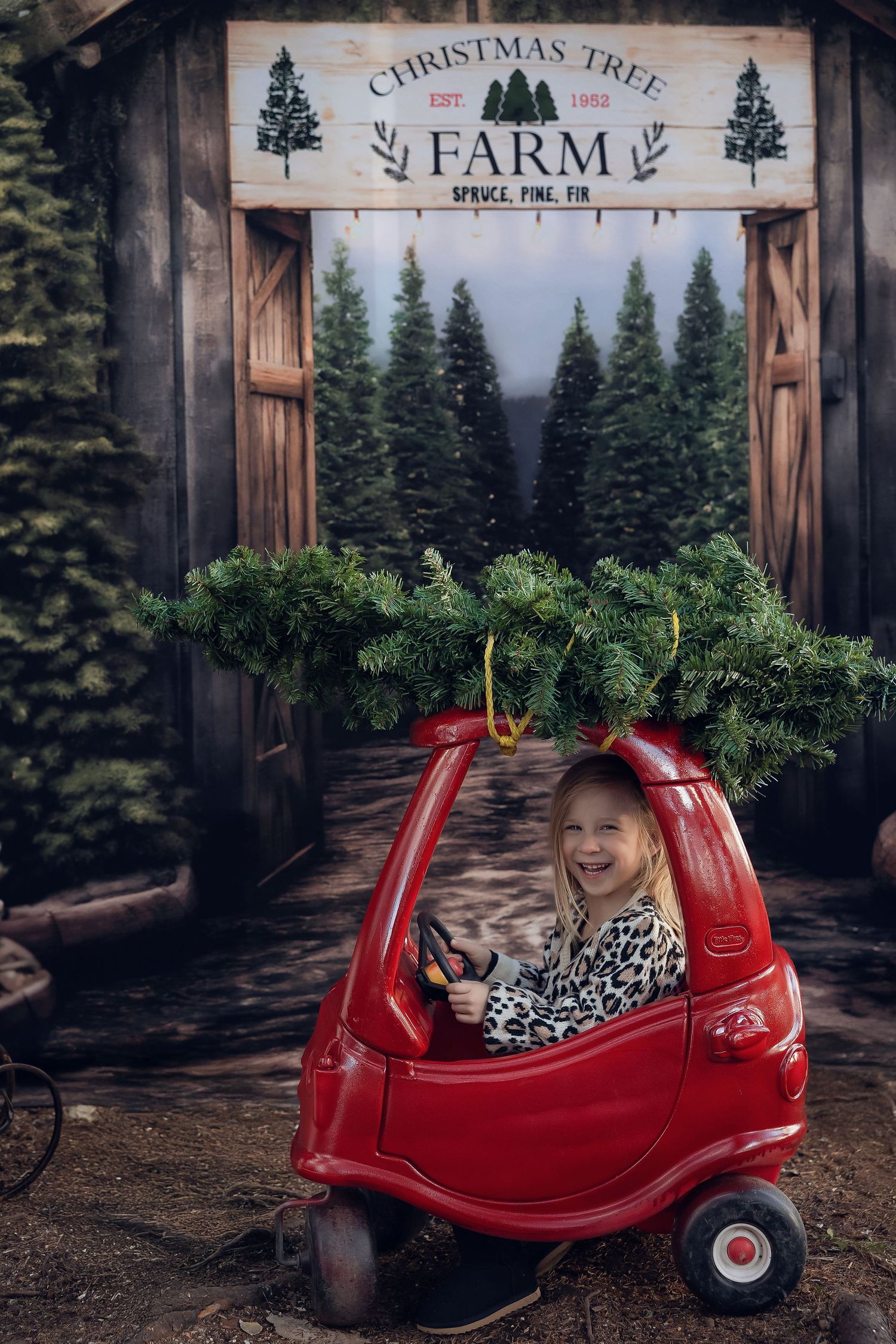 Smiling toddler girl sitting in red toy car with Christmas tree on top at a festive Christmas Tree Farm backdrop.