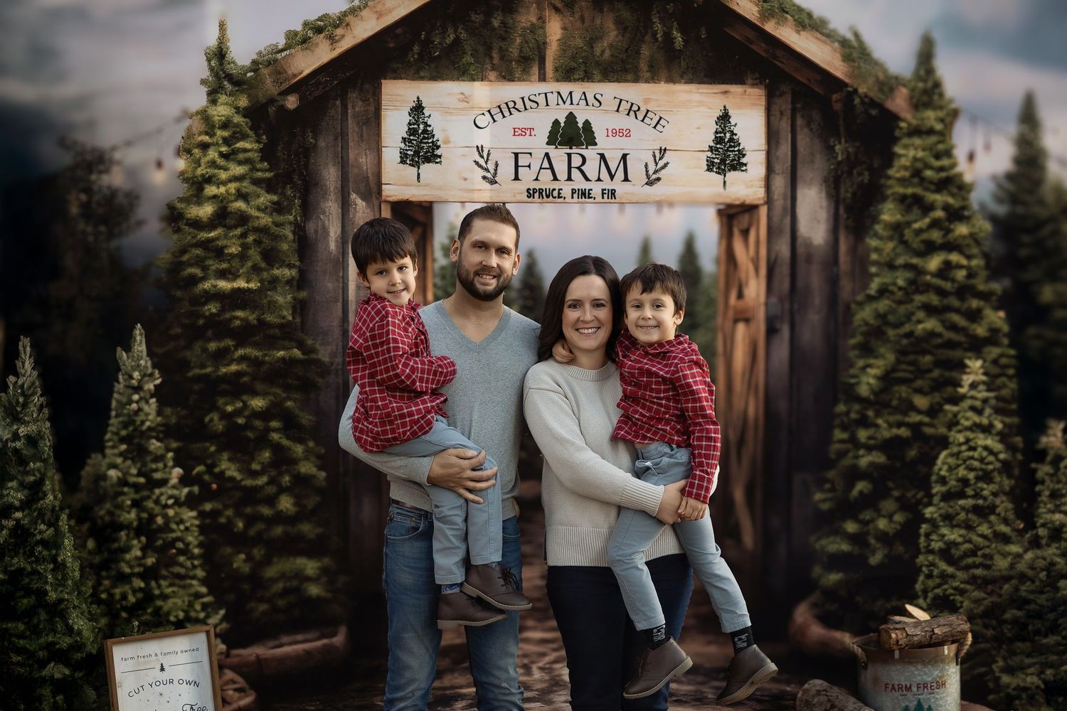 Family of four posing in front of a Christmas Tree Farm backdrop with pine trees and rustic wooden shed.