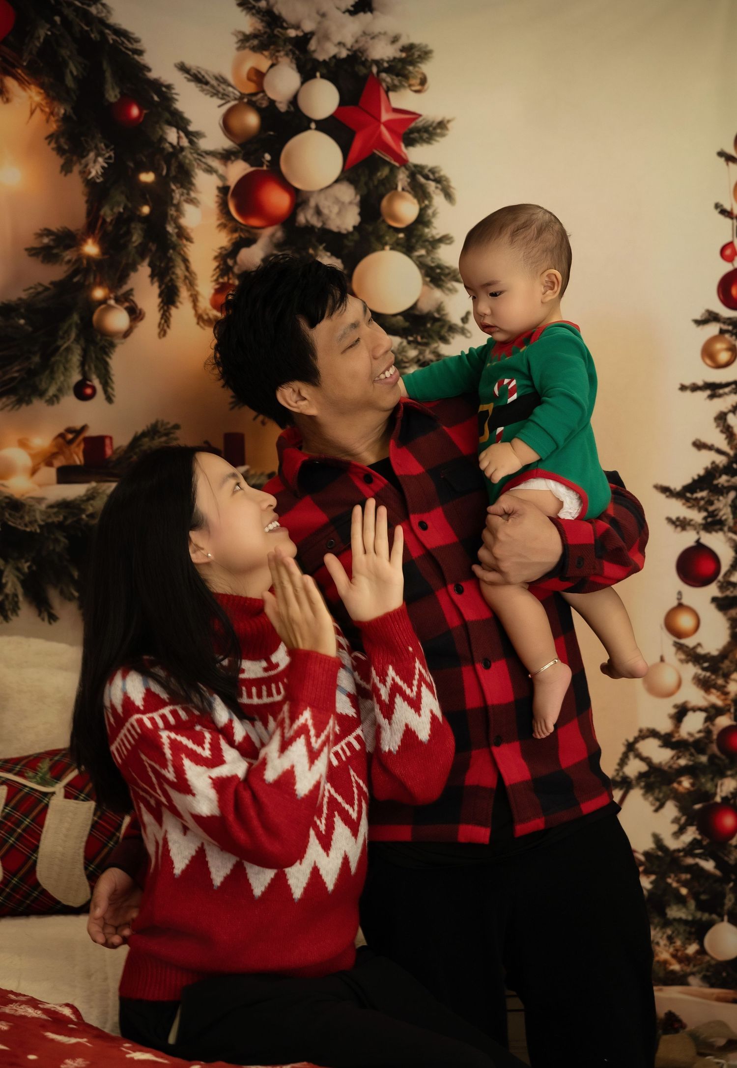 Family in matching red Christmas sweaters joyfully playing with baby near decorated Christmas tree.