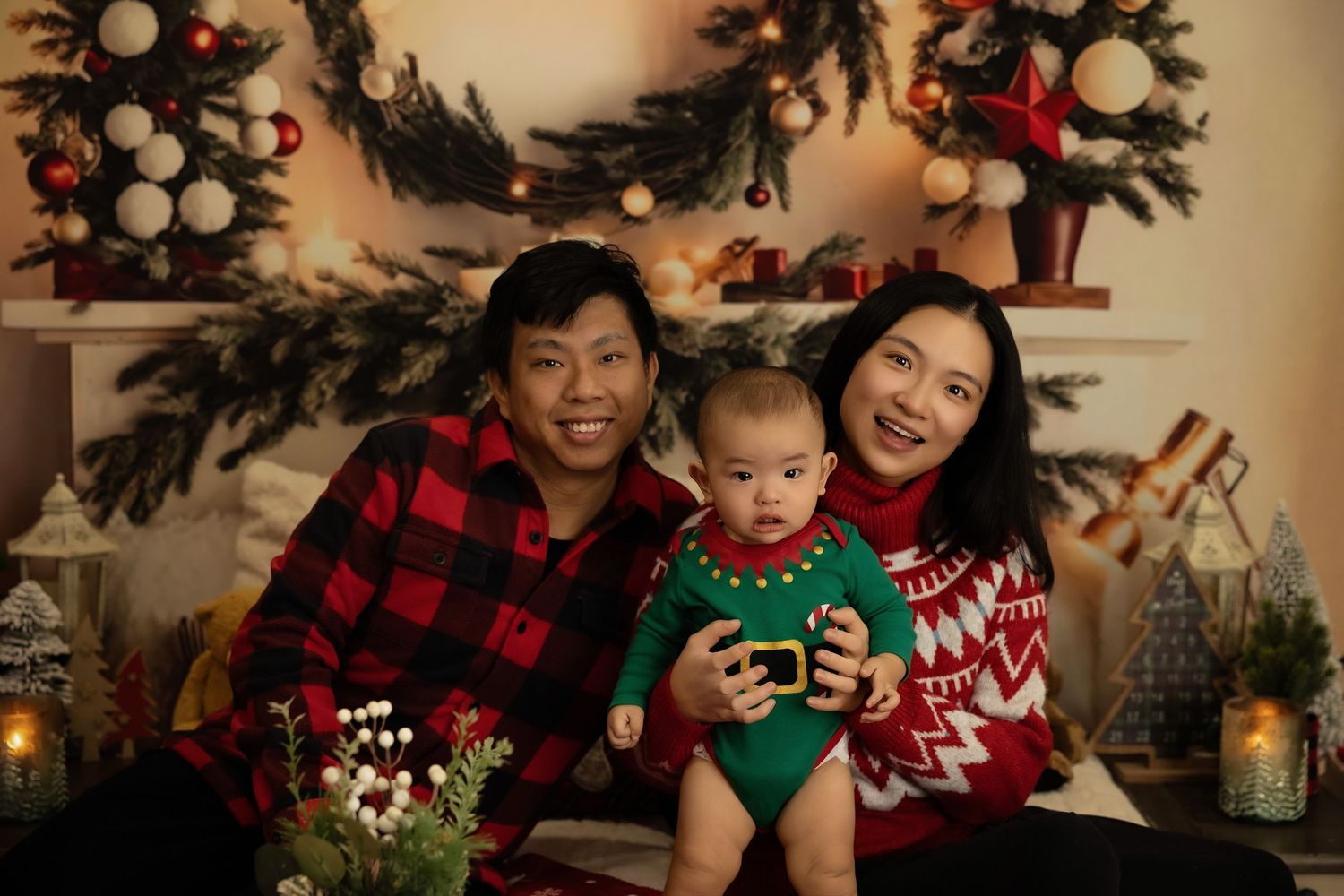 Family Christmas portrait with parents in holiday sweaters holding baby in green outfit, decorated tree backdrop.