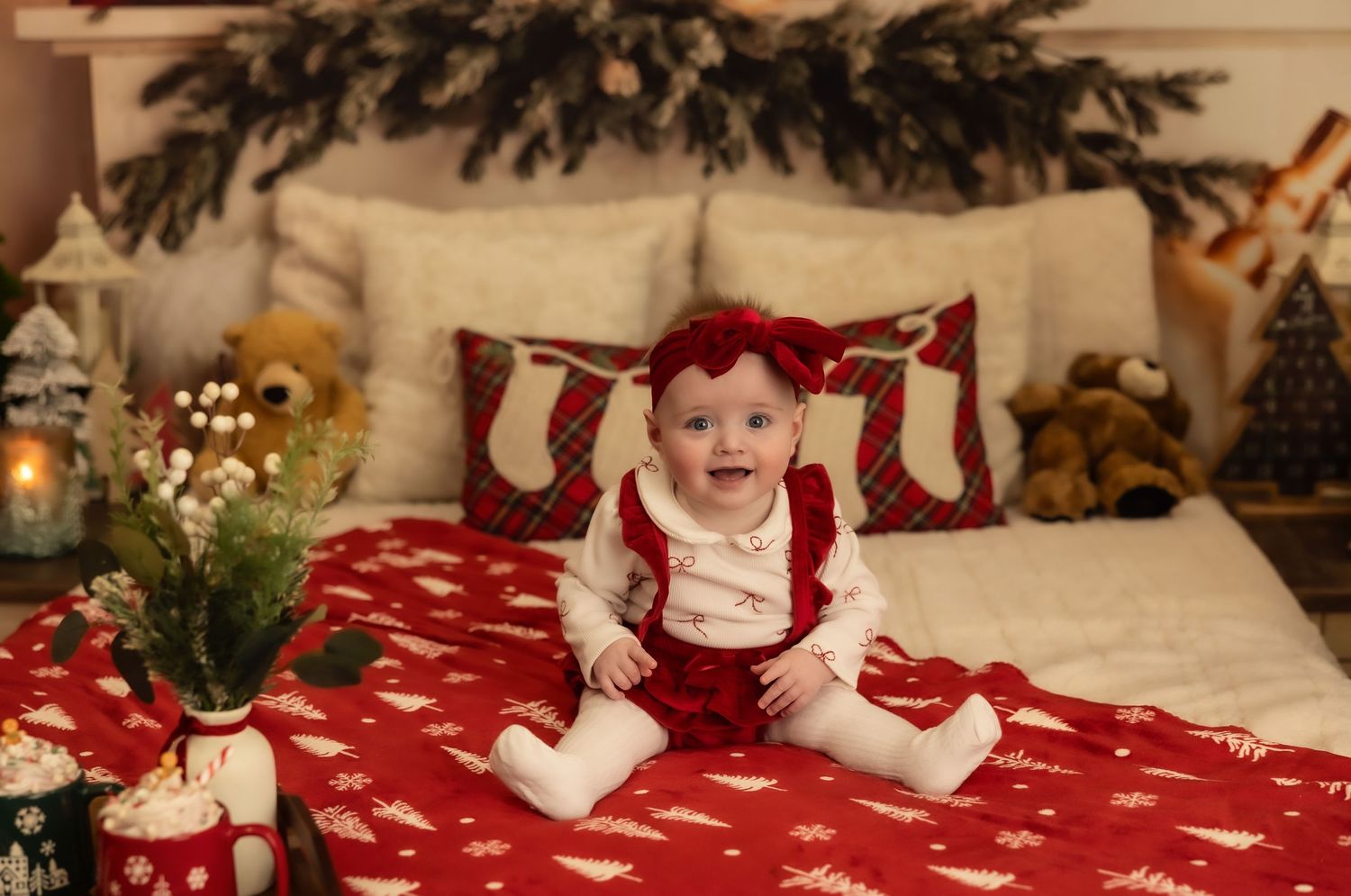 Baby girl in red bow sits on Christmas-decorated bed with snowflake blanket, teddy bears, and holiday greenery.