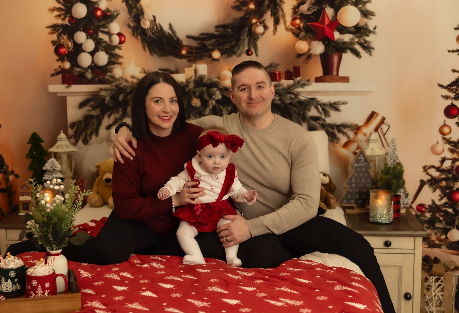 Family Christmas portrait with parents and baby in red outfit sitting on bed with festive decorations and tree.