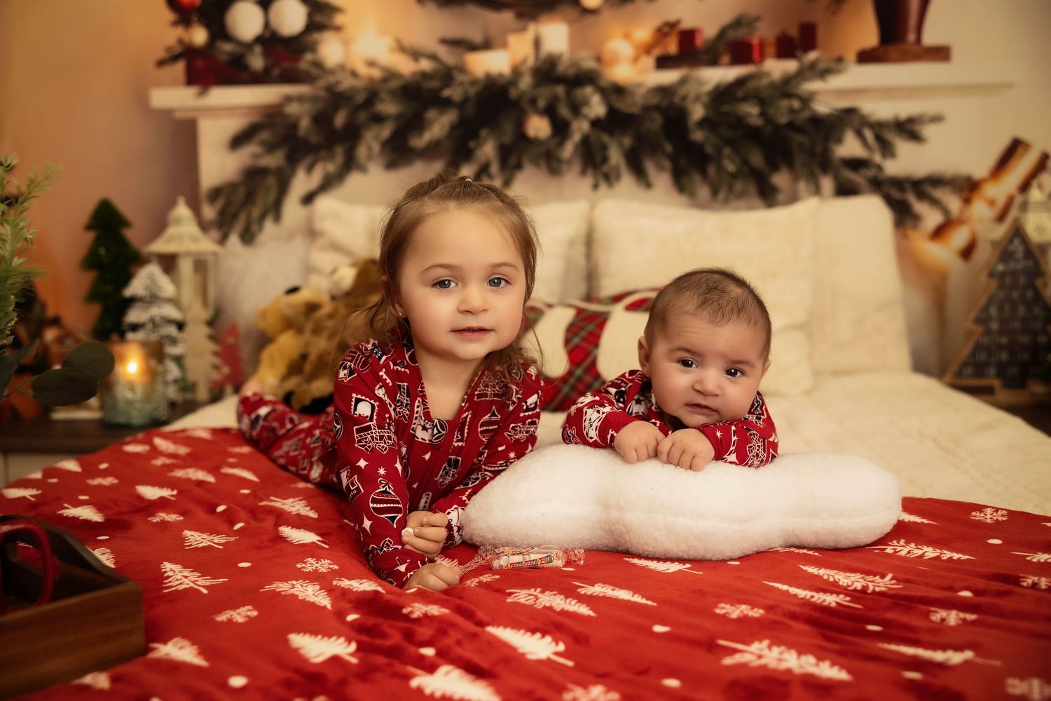 Two children in matching red Christmas pajamas lying on a festive red bed with holiday decorations.