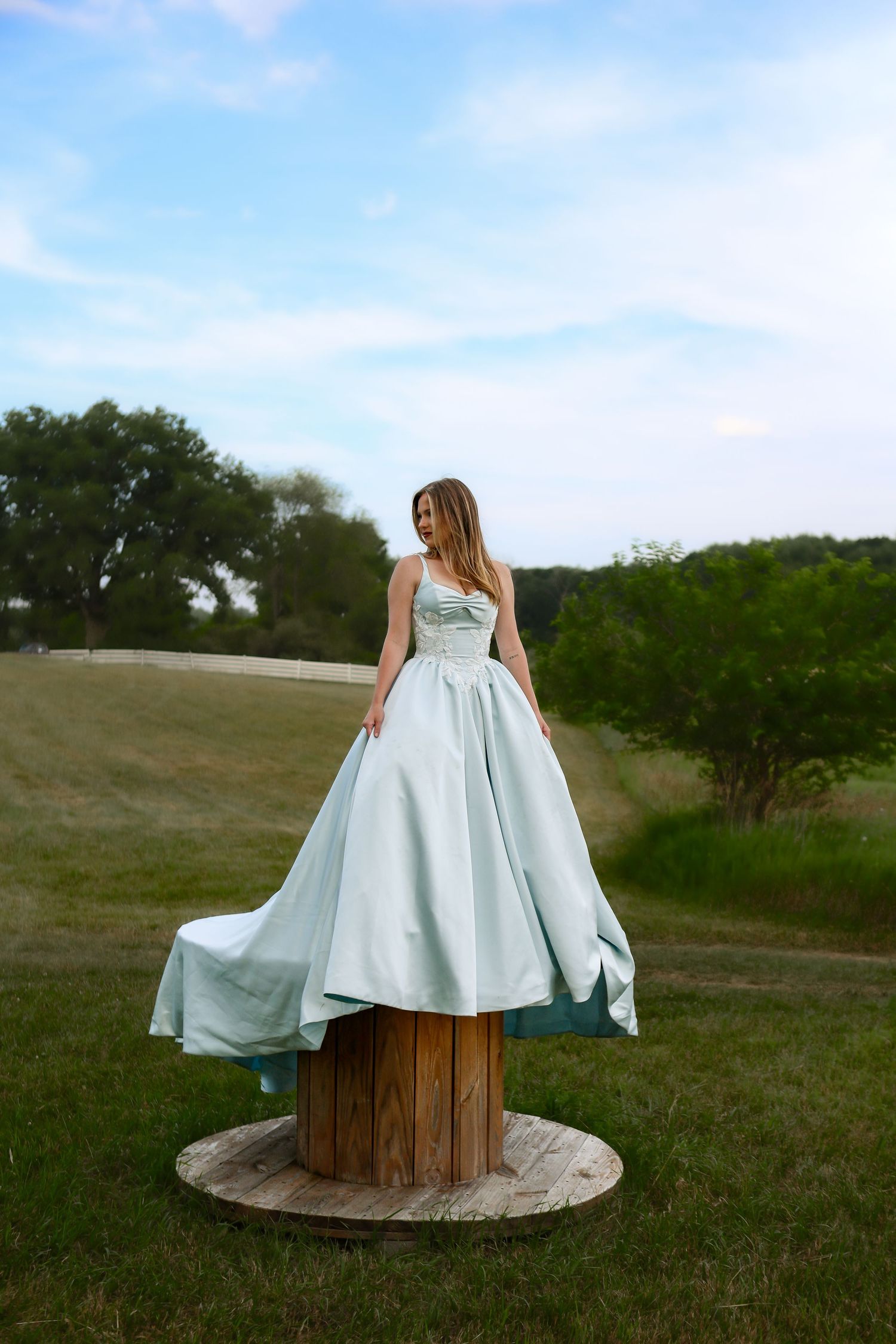 Woman in mint green ball gown standing on wooden spool outdoors with lush green trees and blue sky backdrop.