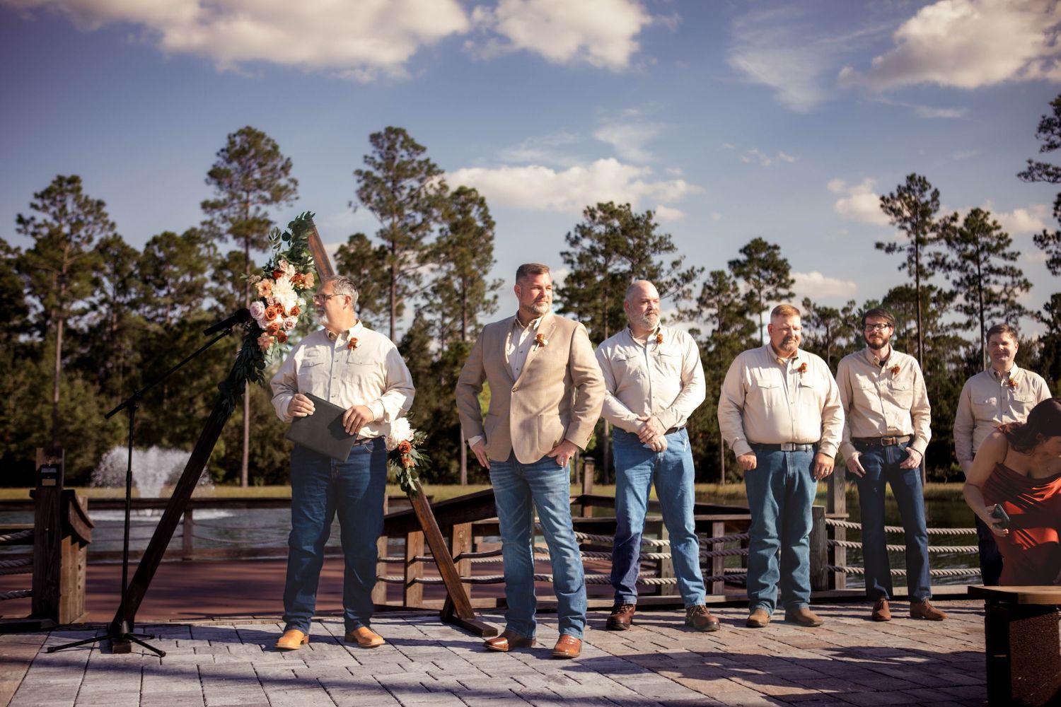 A wedding party wearing khaki and white stand in a row on an outdoor deck with tall pine trees in the background.