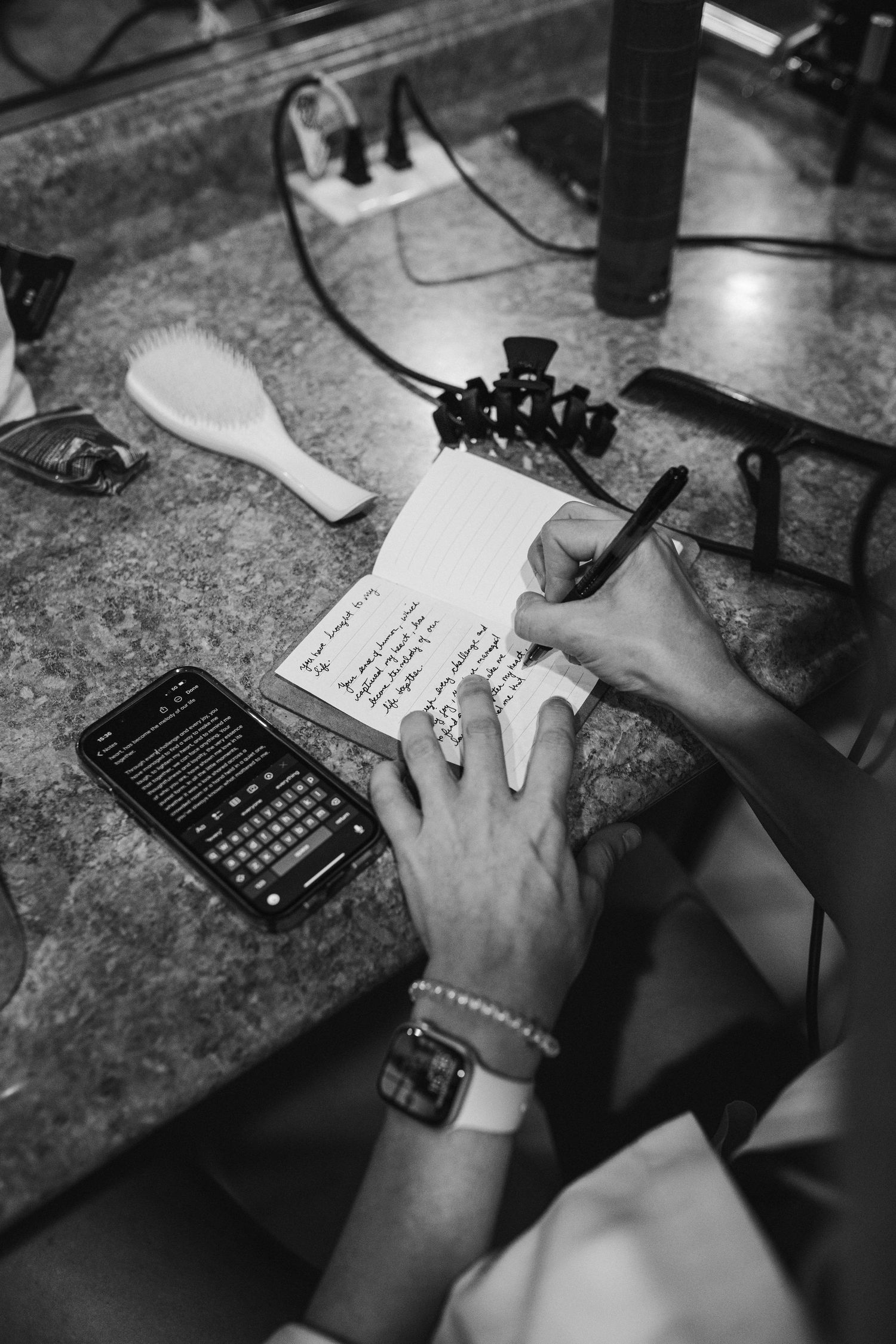 Black and white shot of hands writing on paper with a calculator and charging cable on desk surface.