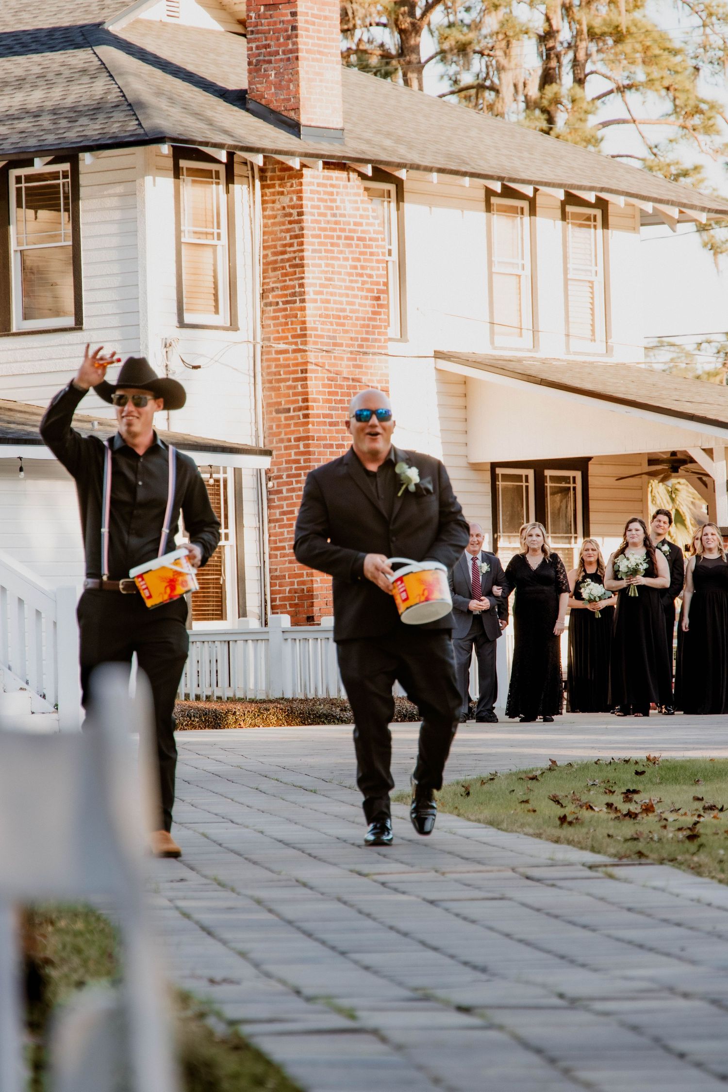 Two people in dark attire carry pumpkins outside a brick house during what appears to be a wedding celebration.