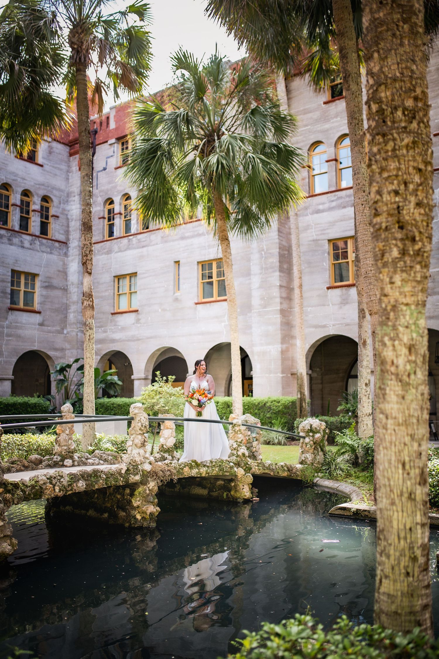 Palm trees line a tranquil courtyard pond surrounded by Spanish colonial architecture with arched windows and stone walls.