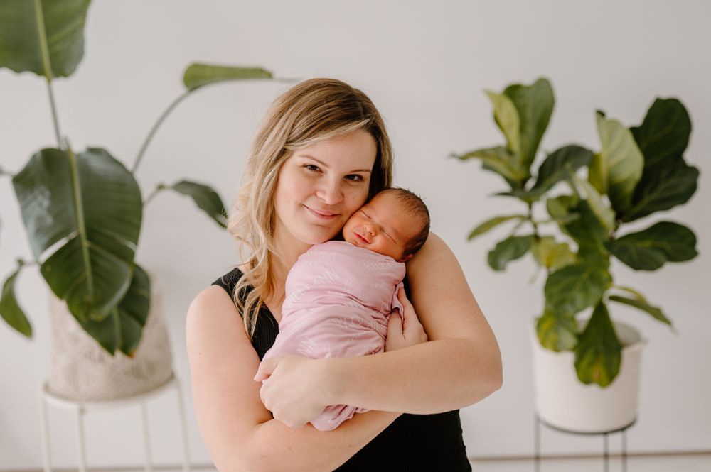 Mother holding sleeping newborn baby in minimalist room with fiddle leaf plants.