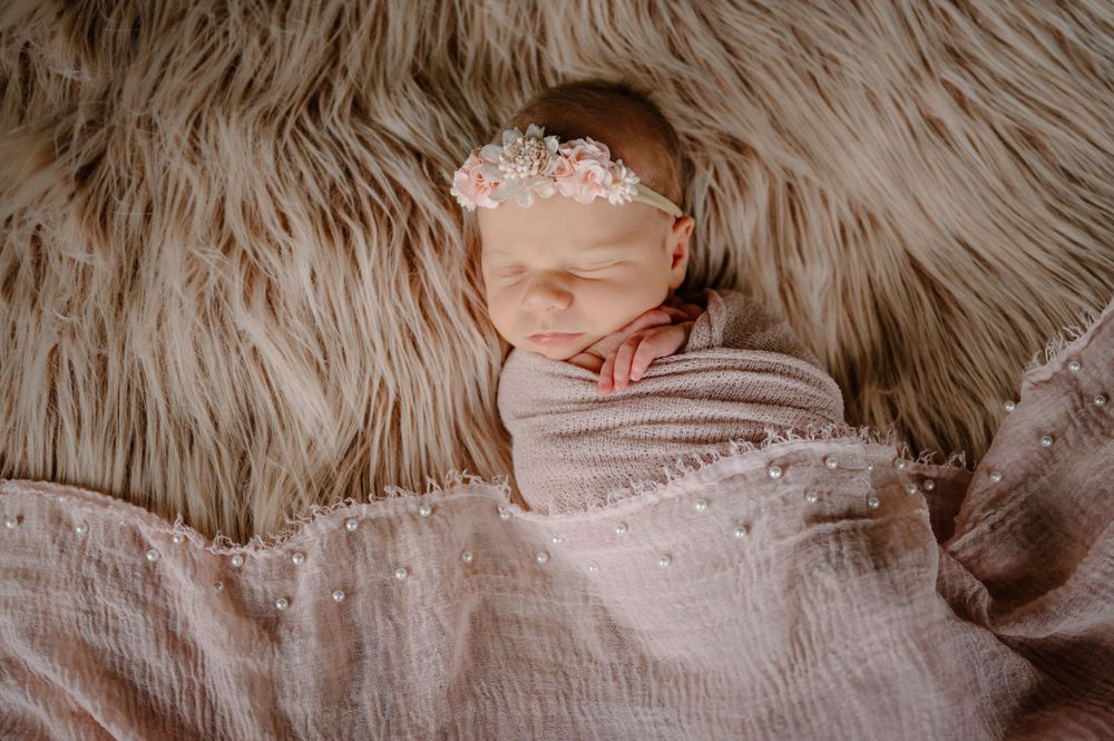 Newborn baby sleeping peacefully on fluffy blanket wearing delicate headpiece.