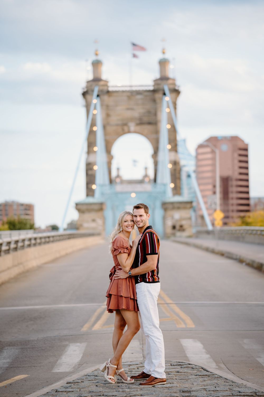 Romantic couple pose on historic bridge with city skyline in background.