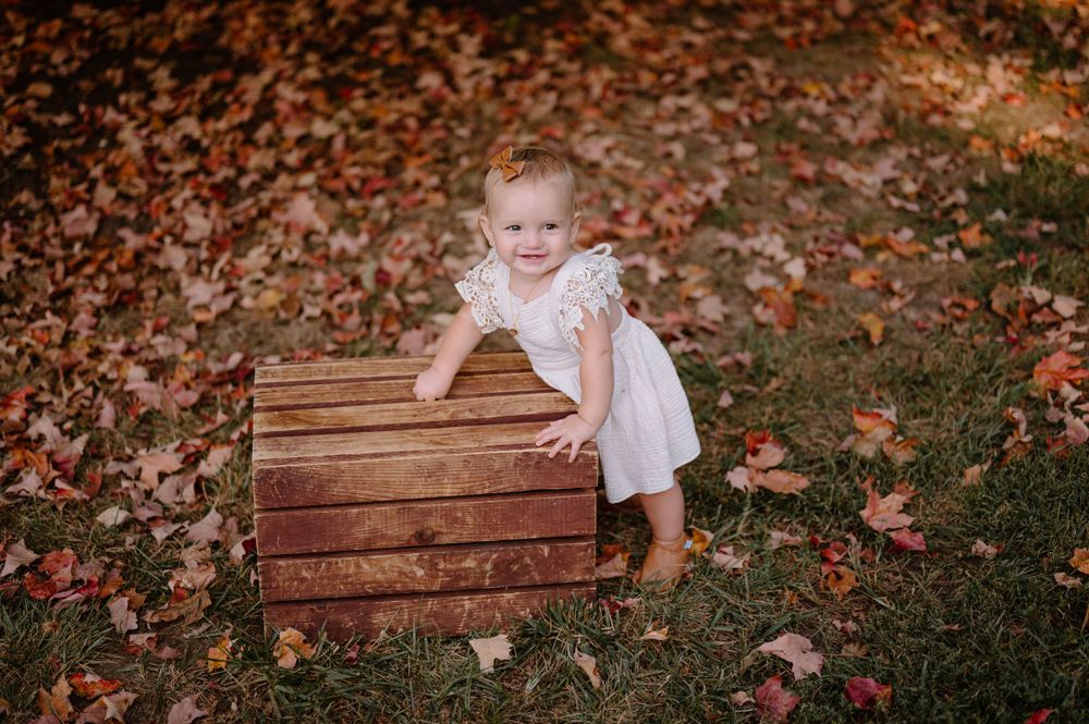 A toddler in a white dress leans against a wooden crate surrounded by autumn leaves.
