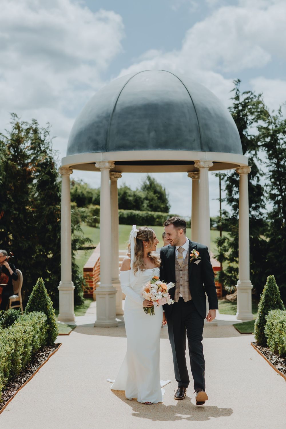 A newlywed couple stands beneath an elegant white domed gazebo surrounded by manicured gardens on their wedding day. The Marquis suffolk