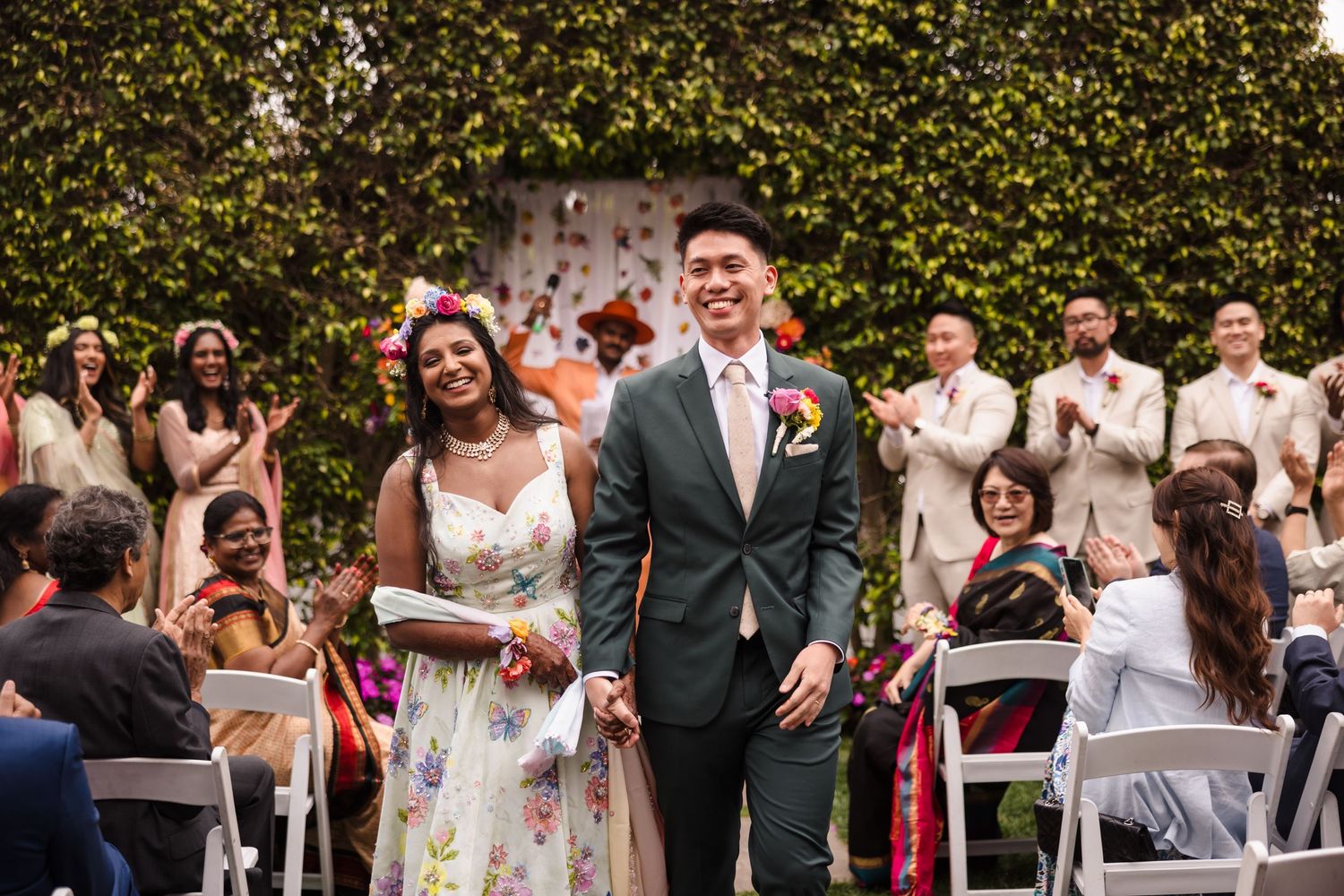 A joyous outdoor wedding celebration with guests seated in white chairs against an ivy wall.