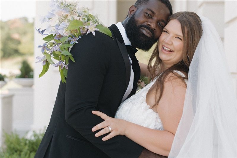 A joyful bride and groom share a laugh together on their wedding day, with white floral decorations nearby.