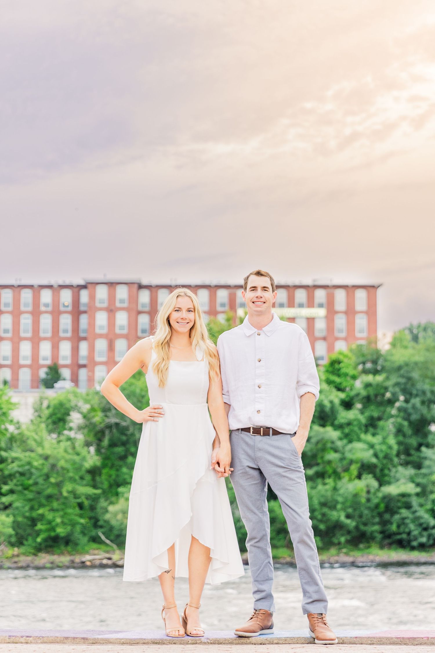 Well-dressed couple stands in front of historic brick mill building at sunset.
