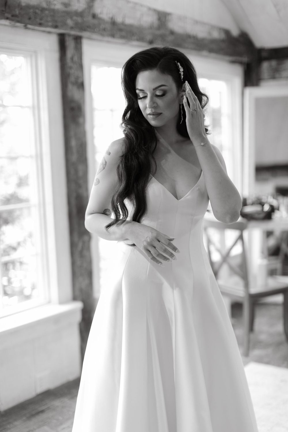 Elegant bride in white gown adjusting hair near rustic window.
