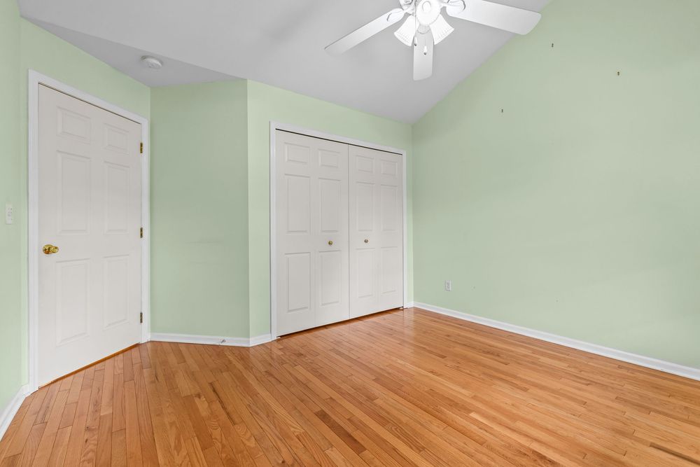 Empty bedroom with light green walls, hardwood floors, ceiling fan, and white closet doors.