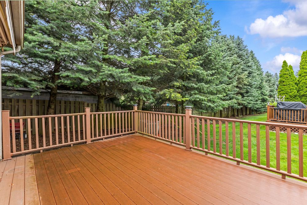 Spacious wooden deck with brown railings overlooking a lush green lawn surrounded by tall evergreen trees.
