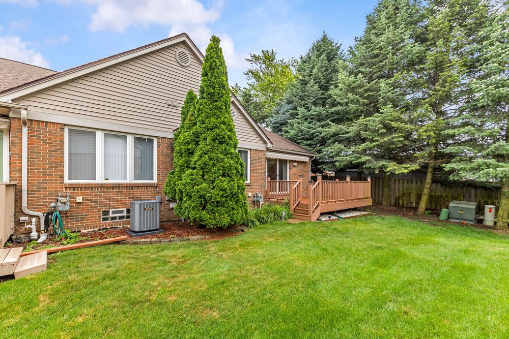 Brick ranch home exterior with wooden deck, central air unit, and lush green yard with evergreen trees.