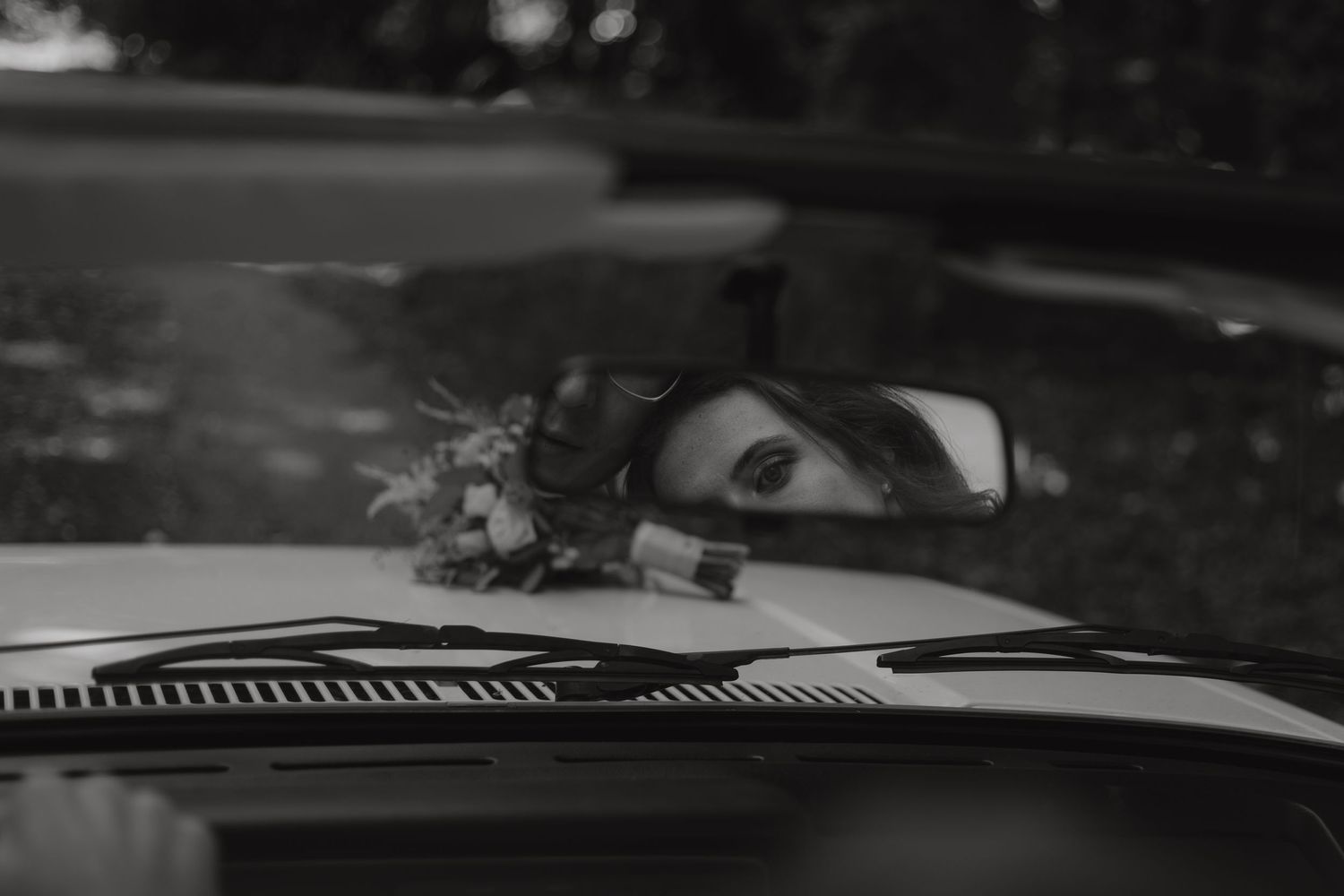 A view through a car windshield in black and white with someone looking in the rearview mirror.
