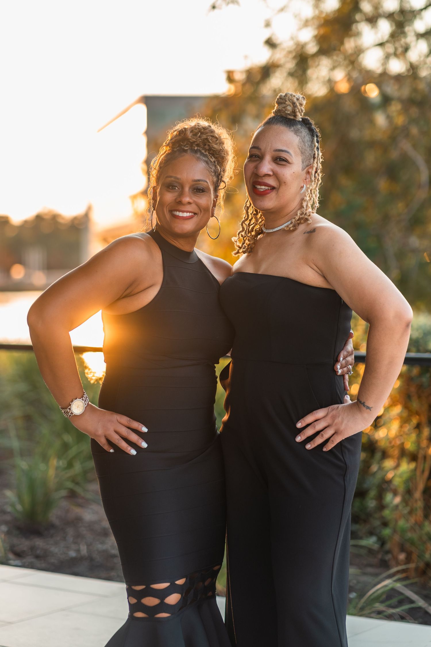 Two friends in black dresses pose together against a golden sunset backdrop by the water.