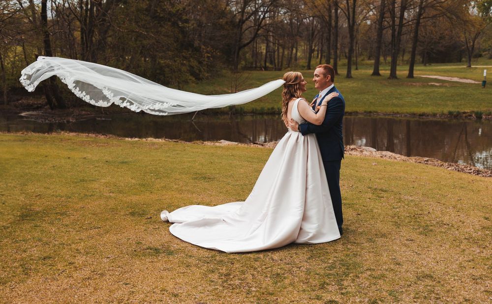 A beautiful wedding scene with a flowing white veil catching the breeze by a lake on an autumn day.