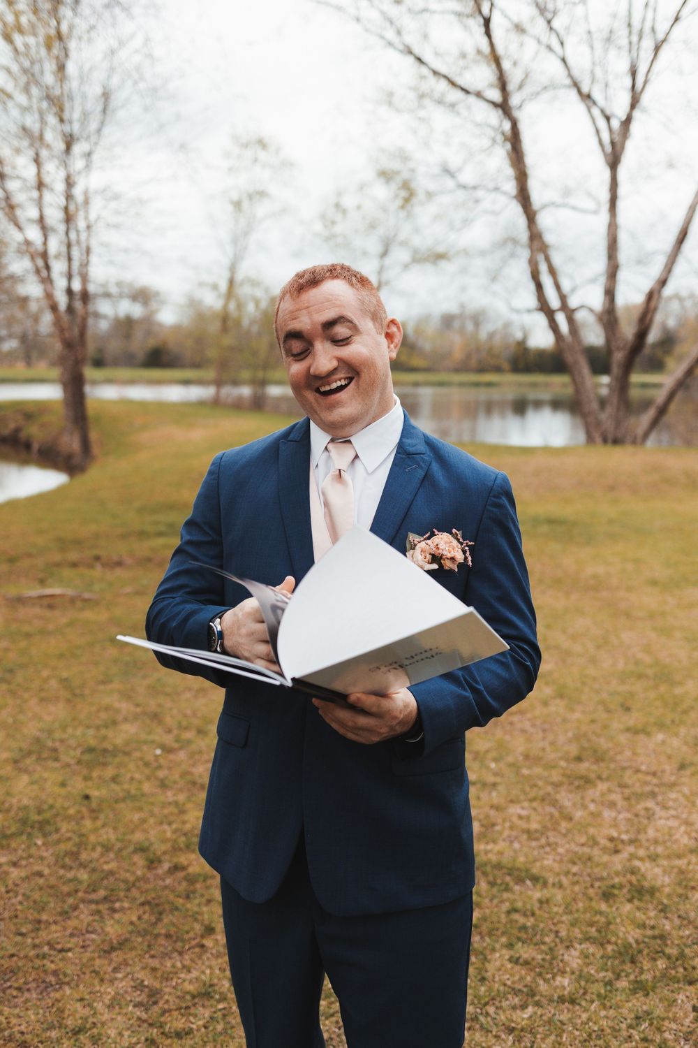 A person in a navy suit excitedly reads from a white notebook outdoors near a pond surrounded by bare trees.