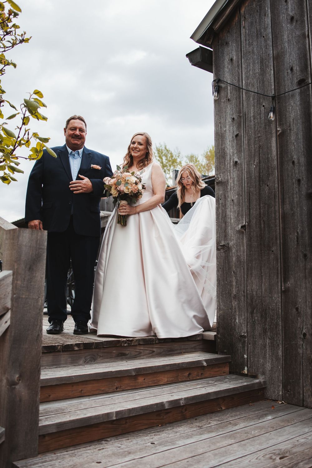 A wedding couple stands on wooden stairs of a rustic barn while the bride holds a bouquet.