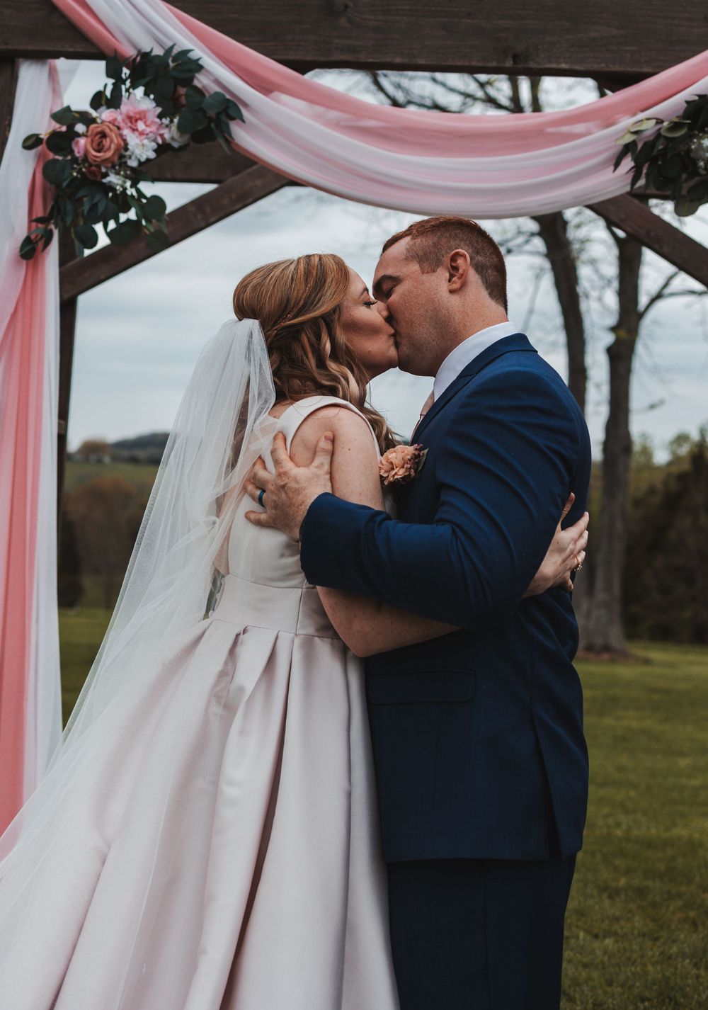 Romantic first kiss sequence under pink draped wedding arch in outdoor garden setting.