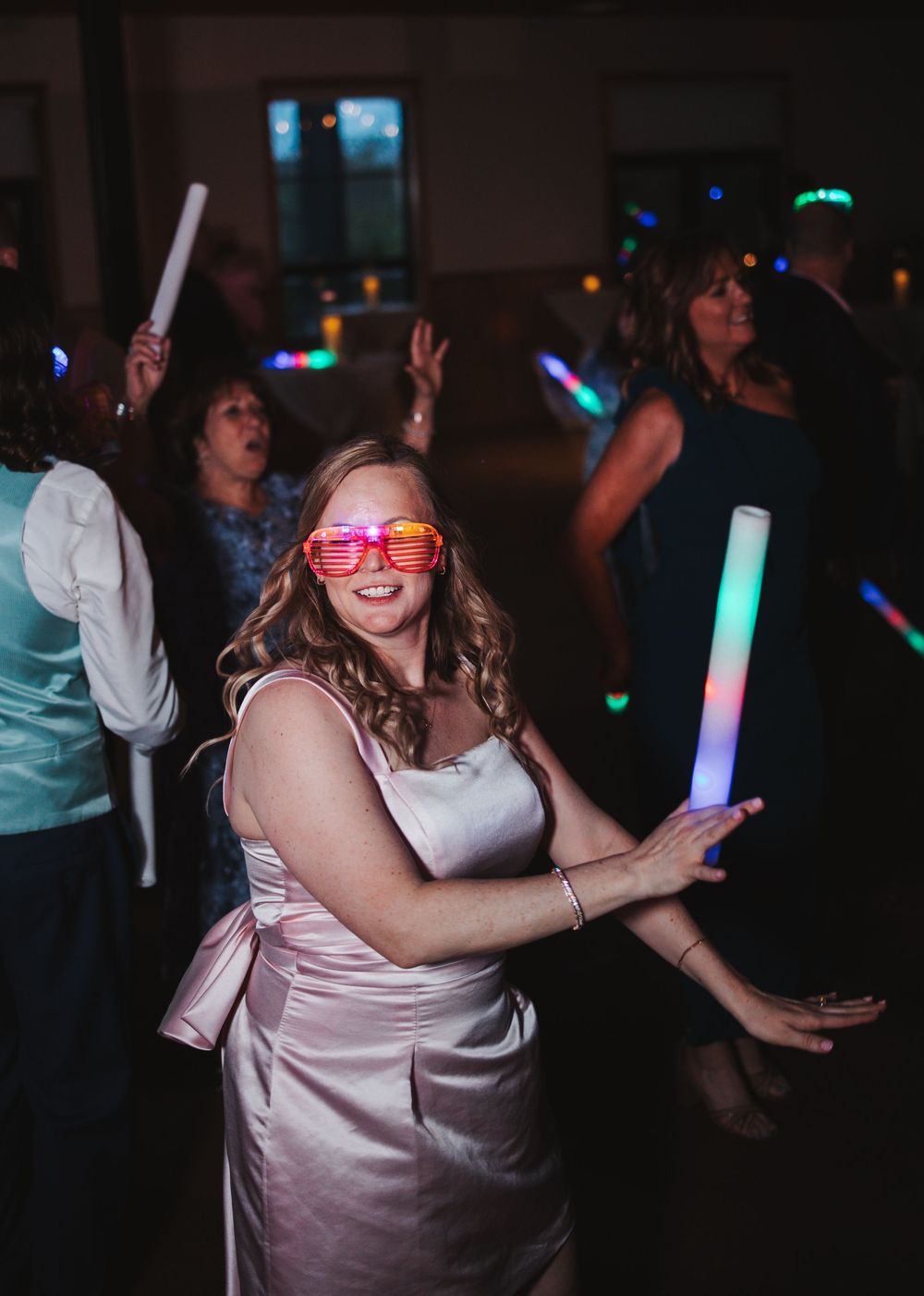 A person in a pink dress dances with glowing LED sticks at a reception with colorful lights in background.