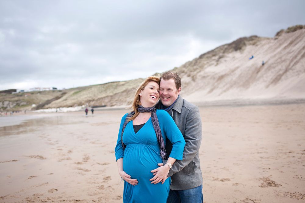 Couple embraces on a sandy beach with cliffs in the background during maternity photoshoot.