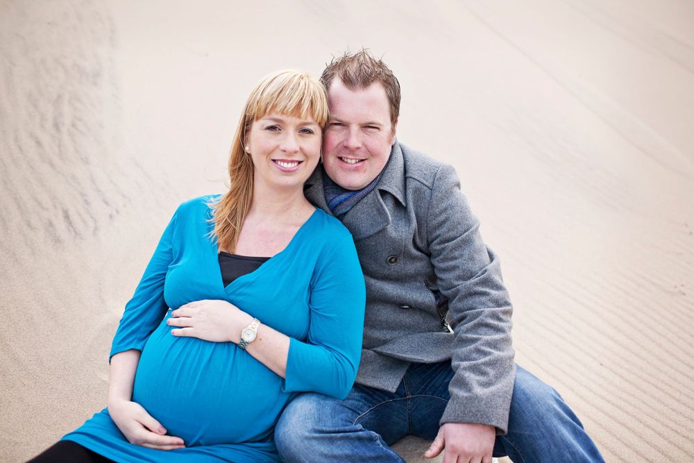 Couple in blue and gray clothing poses lovingly on sandy beach.