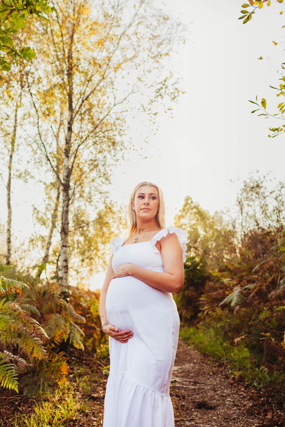 Person in white dress standing on forest path surrounded by autumn foliage and golden sunlight.