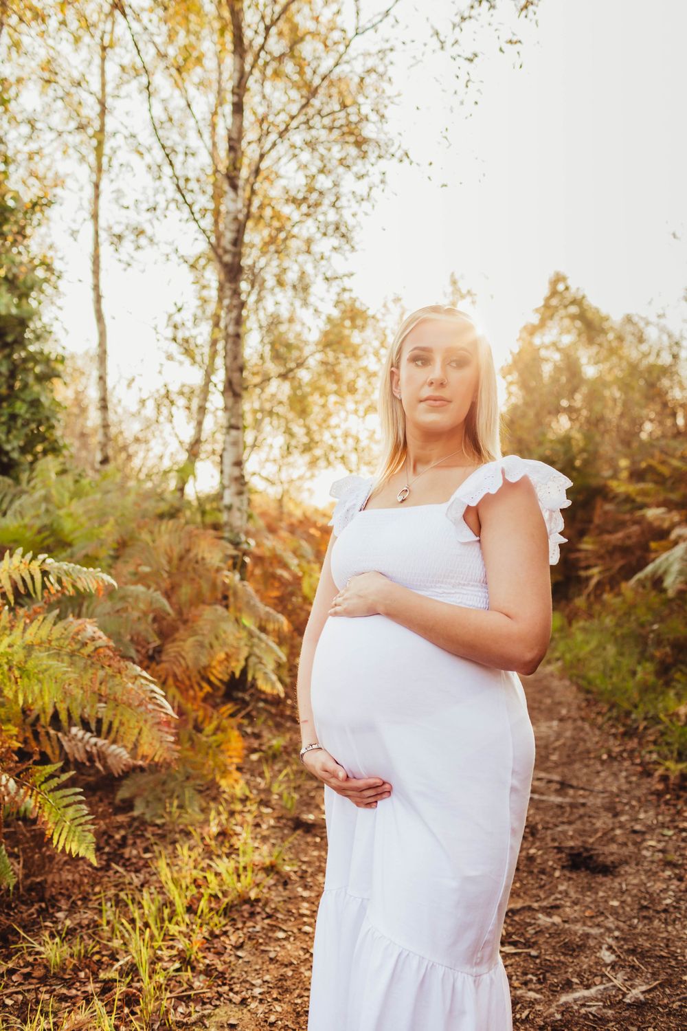 Person in flowing white dress poses on nature trail during golden hour with ferns and trees.