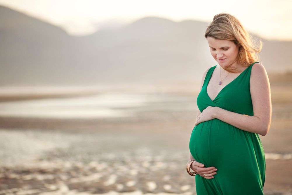 Expectant mother in emerald dress poses on beach at sunset.