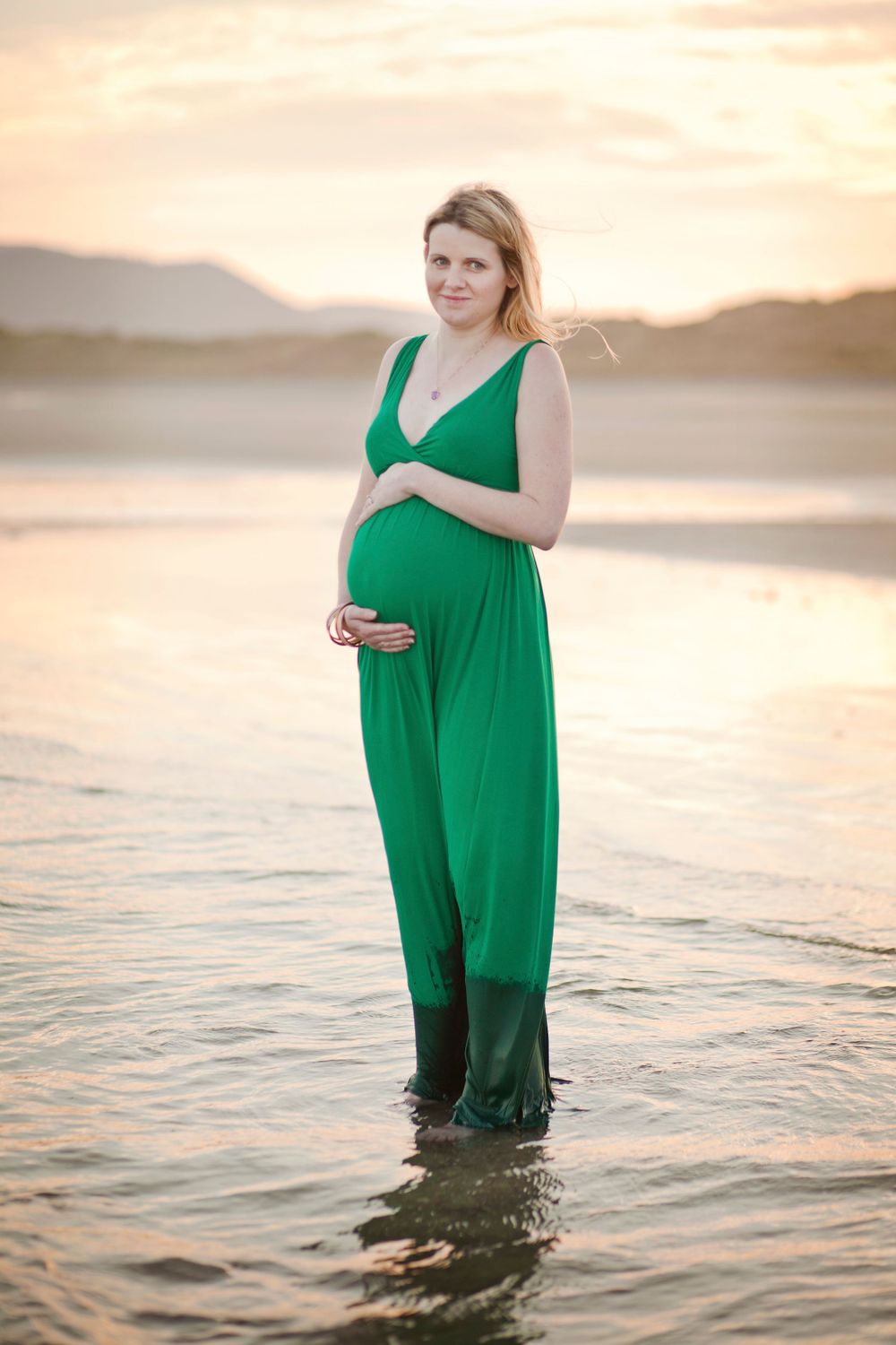 Woman wearing emerald dress stands in shallow ocean water at dusk.