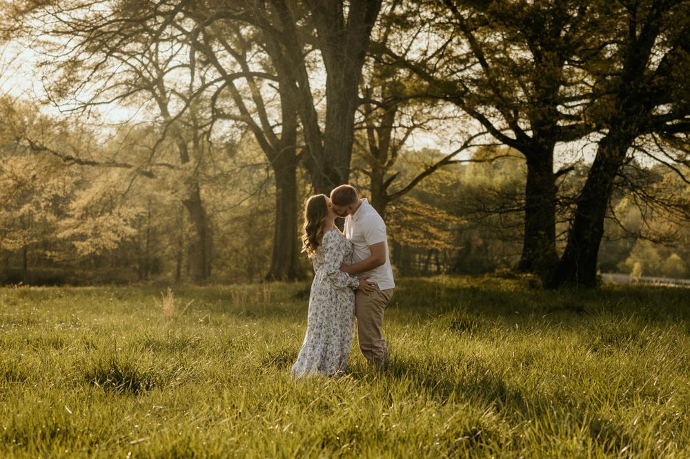Couple in romantic pose standing together in sunlit field with trees in background.
