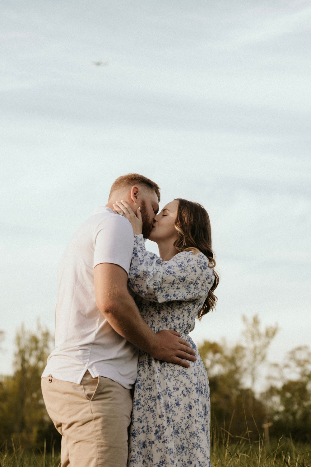 Intimate embrace between couple in casual attire against cloudy sky in field.