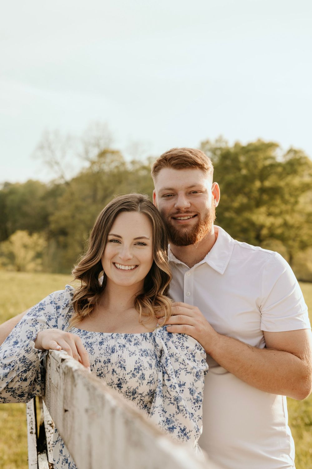 Cheerful couple posing by wooden fence in sunny field wearing coordinated casual outfits.