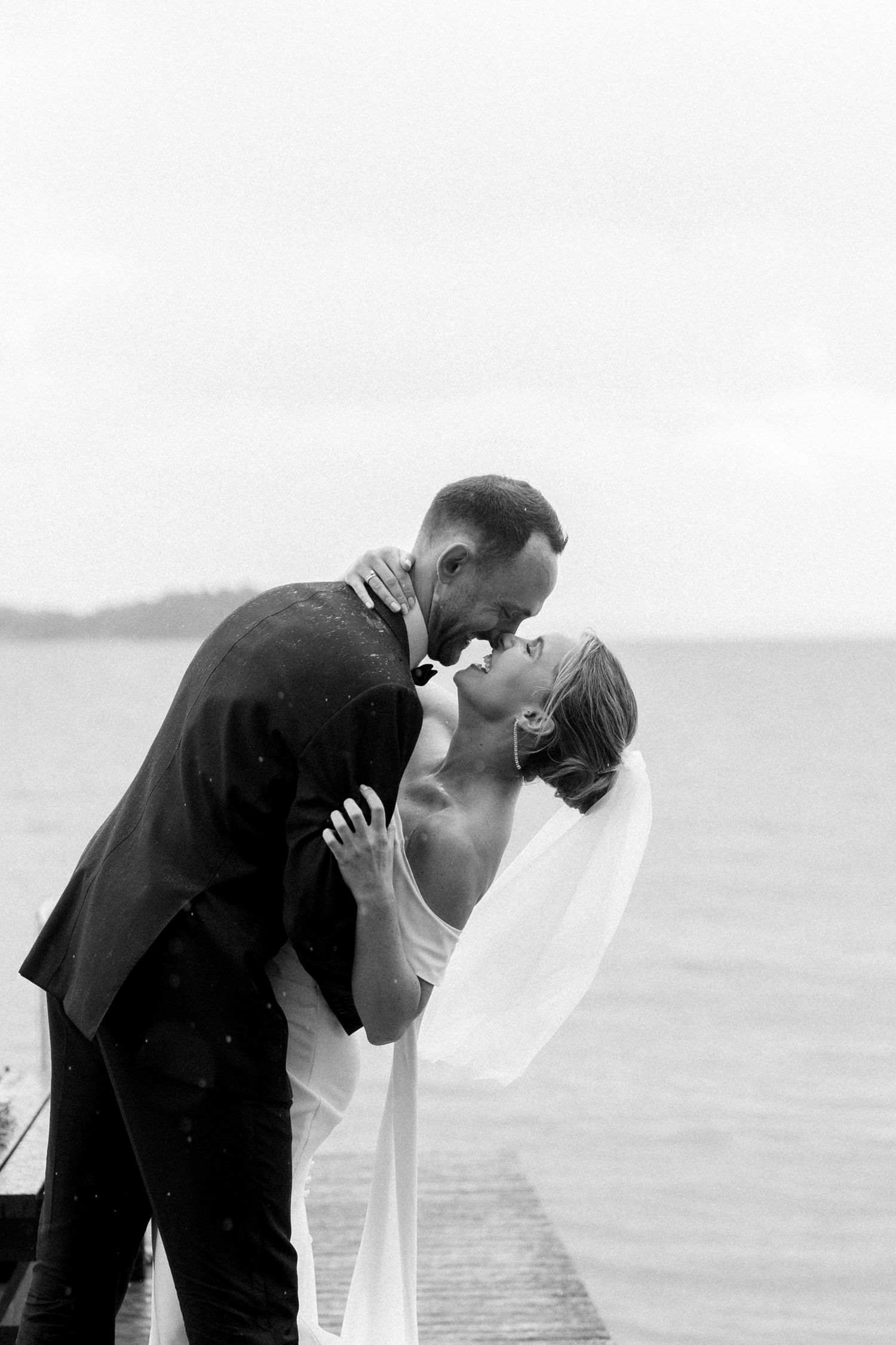 A romantic black and white wedding photo of a couple laughing and kissing in the rain on a dock by the water in Turku, Finland.